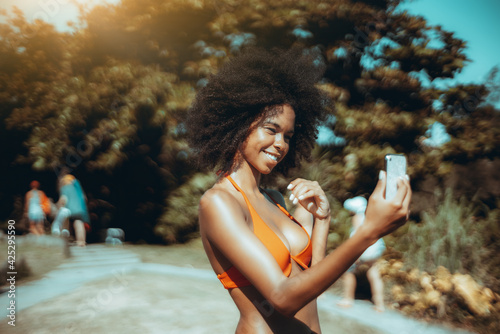 Portrait of laughing young flirtatious African-American female in a swimsuit having video call with her boyfriend via cellphone while standing in a beach area or a tropical resort near a coastal park