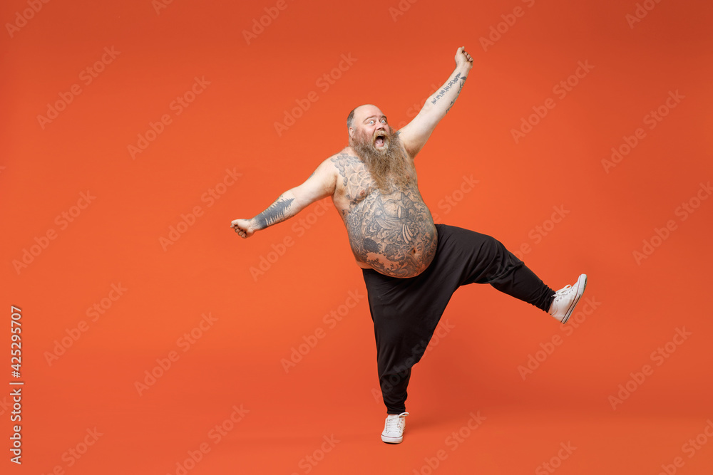 Full length fat overjoyed pudge obese chubby overweight tattooed bearded big belly man in black pants naked torso do winner gesture clench fist outstretch hands isolated on orange background studio.