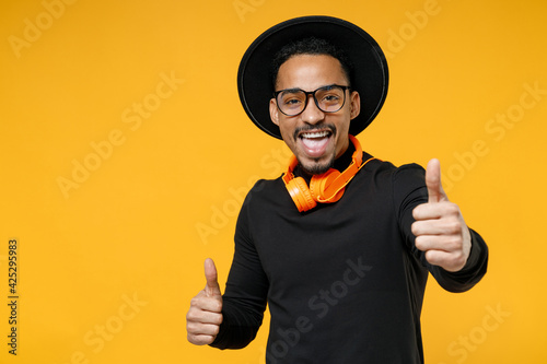 Young music lover happy satisfied smiling african man 20s wear stylish black shirt hat eyeglasses orange headhones showing thumb up like gesture isolated on yellow color background studio portrait photo