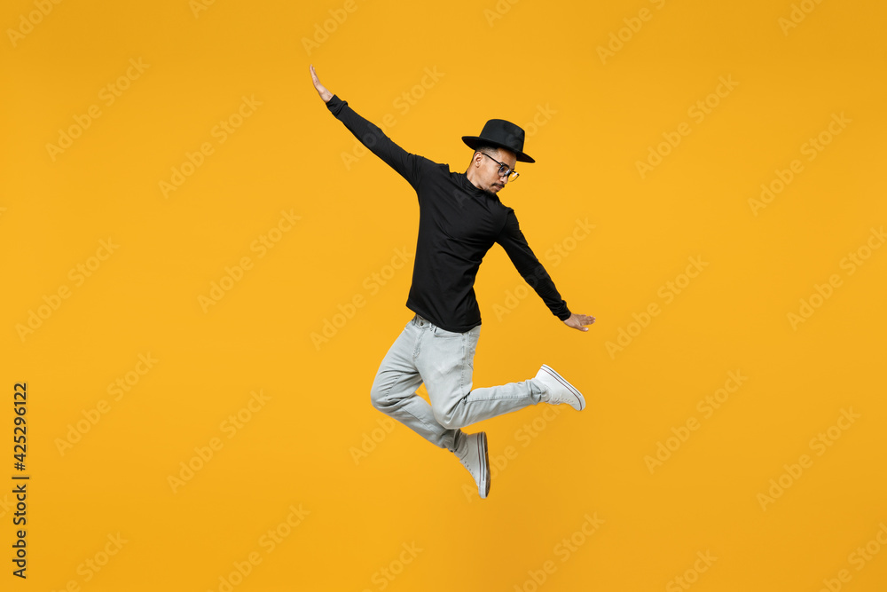 Full length of young active fashionable overjoyed fun happy excited african man in stylish black hat shirt eyeglasses jump high with outstretched hands isolated on yellow background studio portrait