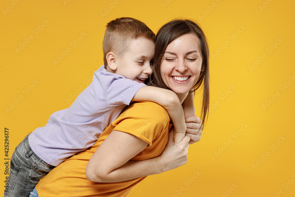 Happy young woman have fun with cute child baby boy 5-6-7 years old in violet t-shirt stand behind hug kiss Mommy little kid son together isolated on yellow background studio Mother's Day love family
