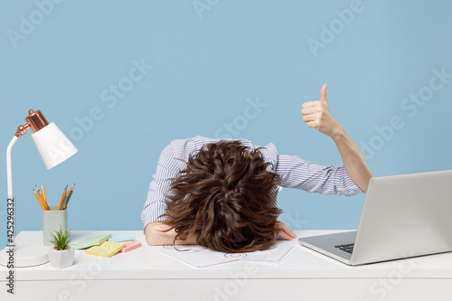 Young tired exhausted secretary employee business woman in shirt sit work sleep laid her head down on white office desk with pc laptop show thumb up gesture isolated on pastel blue background studio.