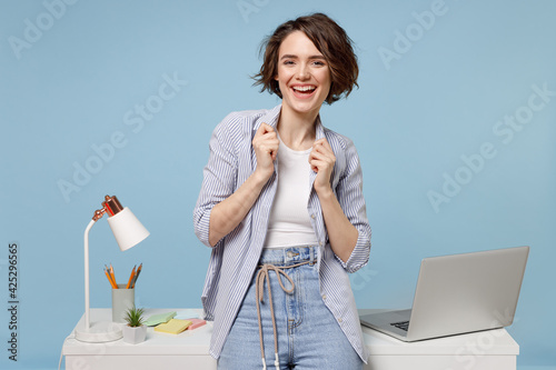Young overjoyed successful employee business woman in casual shirt work stand at white office desk with pc laptop do winner gesture clench fist celebrating isolated on pastel blue background studio