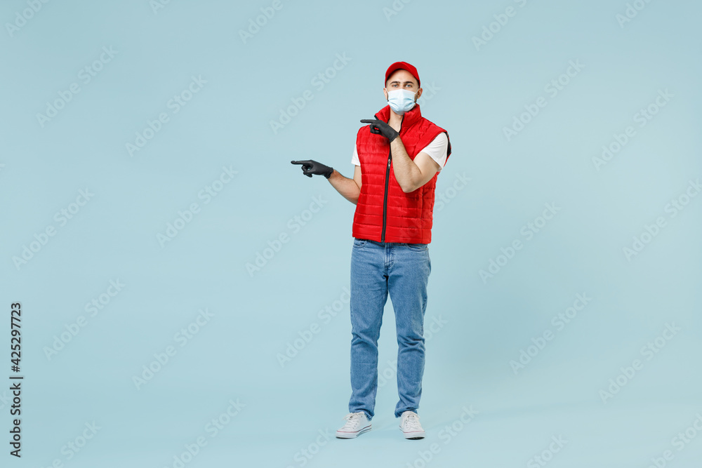 Full length delivery guy employee man in red cap white t-shirt vest uniform sterile face mask gloves work courier service on lockdown covid-19 flu virus stand pose isolated on pastel blue background. Full length delivery guy employee man in red cap white t-shirt vest uniform sterile face mask gloves work courier service on lockdown covid-19 flu virus stand pose isolated on pastel blue background.