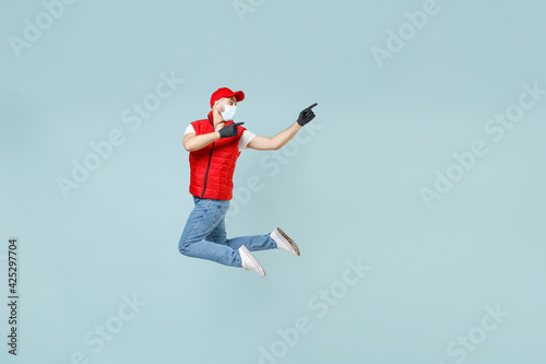 Full length delivery guy employee man in red cap white t-shirt vest uniform sterile face mask gloves work courier service on lockdown covid-19 flu virus run go jump isolated on pastel blue background.