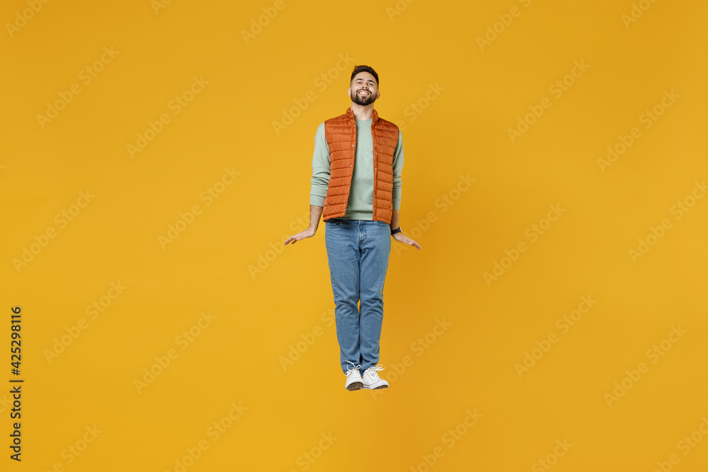 Full length young smiling happy cheerful fun man 20s years old wearing orange vest mint sweatshirt jumping high looking camera isolated on yellow background studio portrait. People lifestyle concept. Full length young smiling happy cheerful fun man 20s years old wearing orange vest mint sweatshirt jumping high looking camera isolated on yellow background studio portrait. People lifestyle concept.