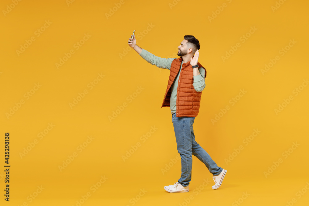 Full length young smiling friendly man 20s in orange vest mint sweatshirt doing selfie shot on mobile phone waving hand greet say hello isolated on yellow background studio. People lifestyle concept. Full length young smiling friendly man 20s in orange vest mint sweatshirt doing selfie shot on mobile phone waving hand greet say hello isolated on yellow background studio. People lifestyle concept.