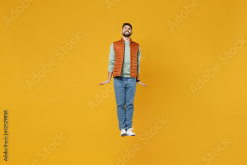 Full length young smiling happy cheerful fun man 20s years old wearing orange vest mint sweatshirt jumping high looking camera isolated on yellow background studio portrait. People lifestyle concept.