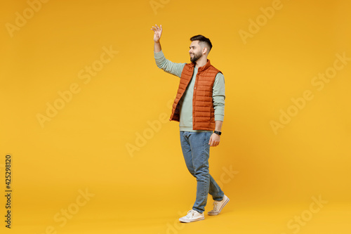 Full length young smiling happy confident smiling cheerful fun caucasian man 20s wearing orange vest mint sweatshirt walking going waving hand greeting isolated on yellow background studio portrait