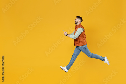 Full length side view young strong powerful fun caucasian man 20s year old in orange vest mint sweatshirt jumping high hold mobile cell phone chat online isolated on yellow background studio portrait © ViDi Studio