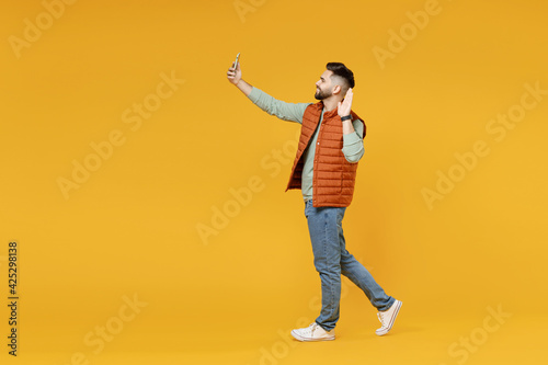 Full length young smiling friendly man 20s in orange vest mint sweatshirt doing selfie shot on mobile phone waving hand greet say hello isolated on yellow background studio. People lifestyle concept.