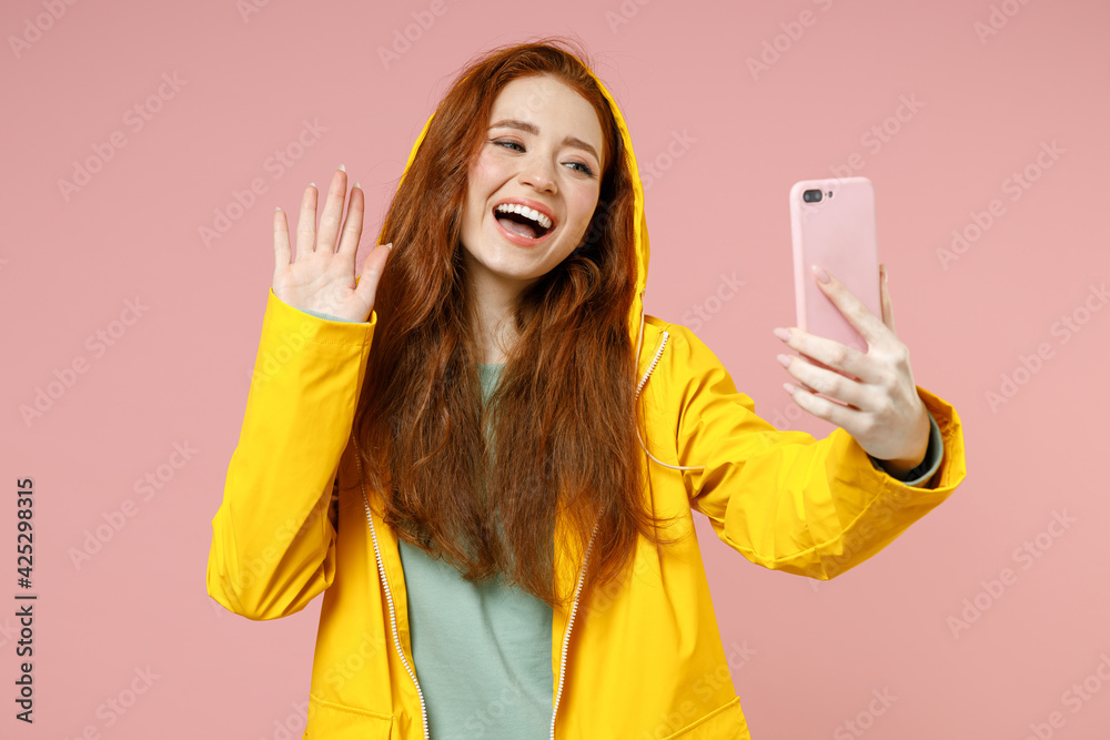Redhead young woman in yellow waterproof raincoat hood outerwear do selfie shot on mobile phone waving hand greet isolated on pastel pink background studio Outdoors lifestyle wet fall season concept Redhead young woman in yellow waterproof raincoat hood outerwear do selfie shot on mobile phone waving hand greet isolated on pastel pink background studio Outdoors lifestyle wet fall season concept