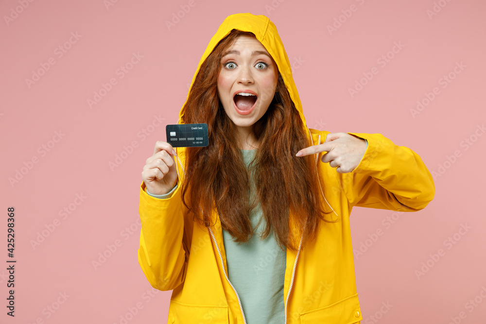Redhead young excited woman 20s in yellow waterproof raincoat hood outerwear point finger on credit bank card isolated on pastel pink background studio. Outdoors lifestyle fall weather season concept