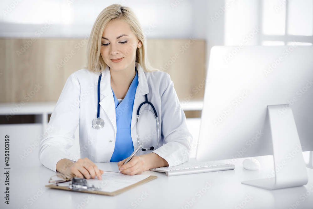 Woman-doctor at work while sitting at the desk in hospital or clinic. Blonde cheerful physician filling up medication history record form Woman-doctor at work while sitting at the desk in hospital or clinic. Blonde cheerful physician filling up medication history record form