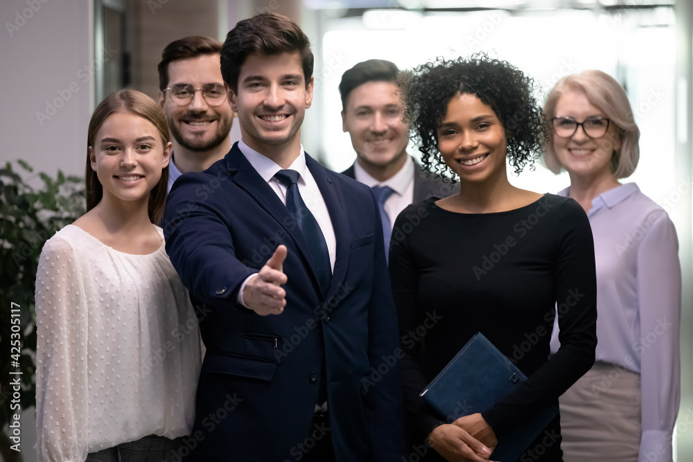 Obraz premium Happy diverse employees with young male leader or boss in front meet welcome new worker into team. Smiling businessman stretch hand for handshake with newcomer in office. Recruitment concept.
