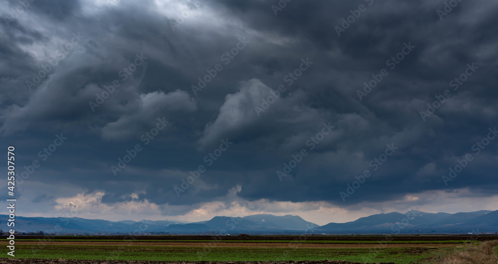 Obraz premium Dramatic storm clouds over empty green agricultural fields, blue mountains at the horizon line.