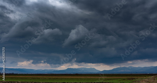 Dramatic storm clouds over empty green agricultural fields, blue mountains at the horizon line. © Alpar
