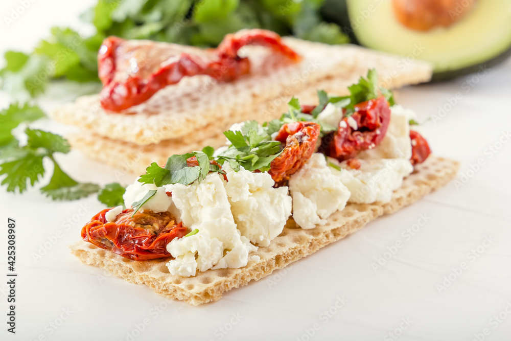 Crispy low-calorie wheat crackers with curd cheese and sun-dried tomatoes on a background of avocado and greens on a white wooden table. Close-up