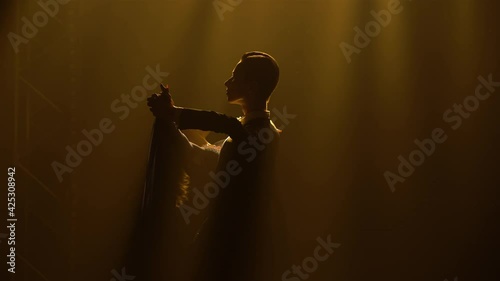 Light waltz movements performed by charming pair of ballroom dancers. Man in an elegant suit and woman in long dress waltz in dark studio with yellow light. Silhouettes. Close up. photo