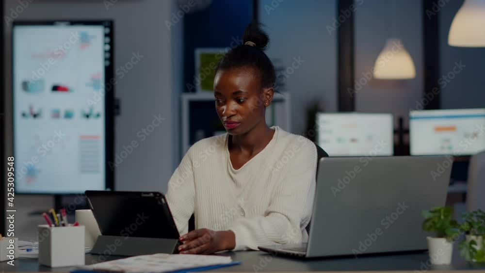 African manager woman using laptop and tablet in same time working overtime in business start-up office. Busy multitasking Busy employee analysing financial statistics overworking writing, searching. African manager woman using laptop and tablet in same time working overtime in business start-up office. Busy multitasking Busy employee analysing financial statistics overworking writing, searching.
