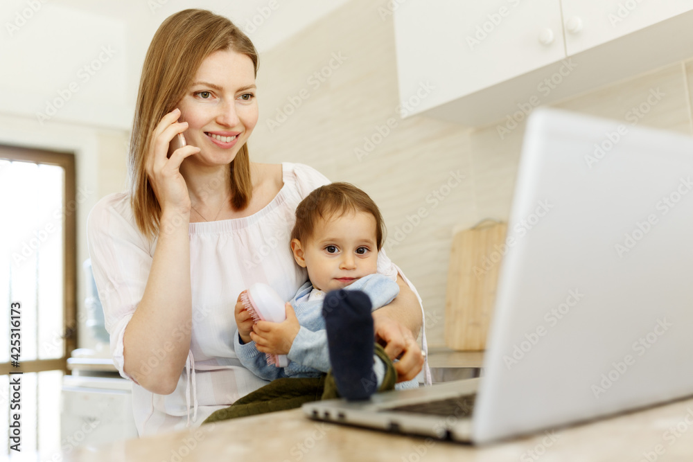 Beautiful business mom is talking on the mobile phone and taking notes while spending time with her cute baby boy at kitchen home office. Working mother concept,Laptop on table Beautiful business mom is talking on the mobile phone and taking notes while spending time with her cute baby boy at kitchen home office. Working mother concept,Laptop on table