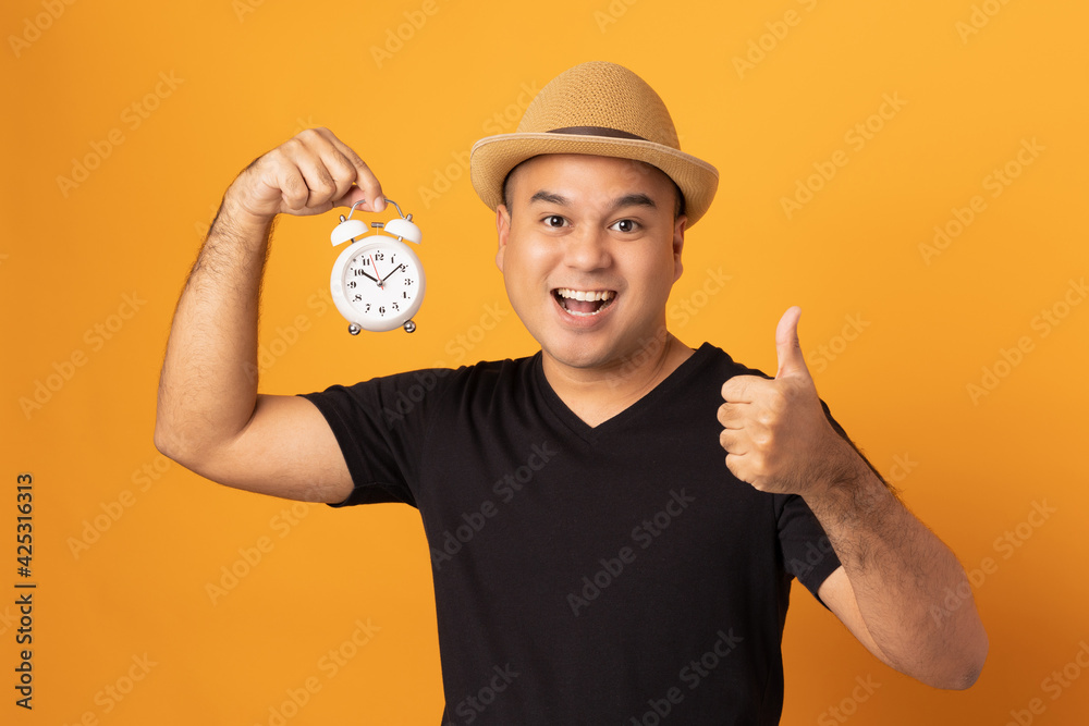 Young asian man wearing hat black shirt holding small alarm clock standing on isolated yellow background. He feeling happy and excited