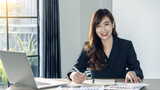 Young Asian businesswoman happily working on documents, graphs and laptops, smiling in the office.