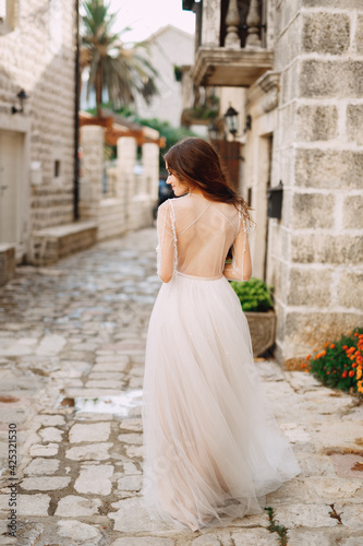 A bride in a stylish dress with an open back stands in front of an ancient building on a cozy Perast street, back view