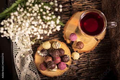 Teezeit in rustikal Still. Schleier Kraut liegt als Deko auf dem Holztisch. Glas  Tee steht auf dem Holzhintergrund, bunte Pralinen liegen liegen auf dem Stück Holz