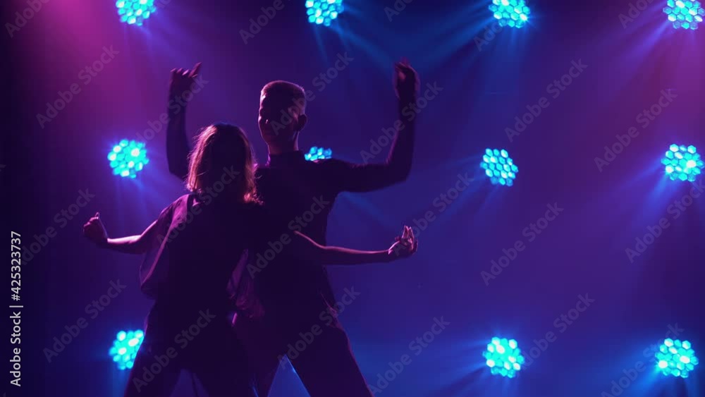 A couple of young dancers are dancing passionate Latin American dance. Rumba, samba, cha-cha-cha, jive. Silhouettes of two dancers on a dark studio smoky background with blue-violet lights. Close up. A couple of young dancers are dancing passionate Latin American dance. Rumba, samba, cha-cha-cha, jive. Silhouettes of two dancers on a dark studio smoky background with blue-violet lights. Close up.