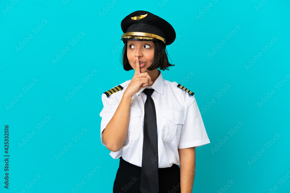 Young airplane pilot over isolated blue background showing a sign of silence gesture putting finger in mouth Young airplane pilot over isolated blue background showing a sign of silence gesture putting finger in mouth