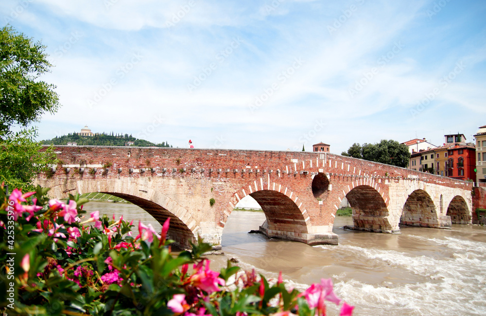 Bridge Ponte Pietra in Verona on Adige river. Veneto region. Italy. Sunny summer day panorama and blue sky with clouds. Santuario della Madonna di Lourdes (Our Lady of Lourdes sanctuary) on the hills 