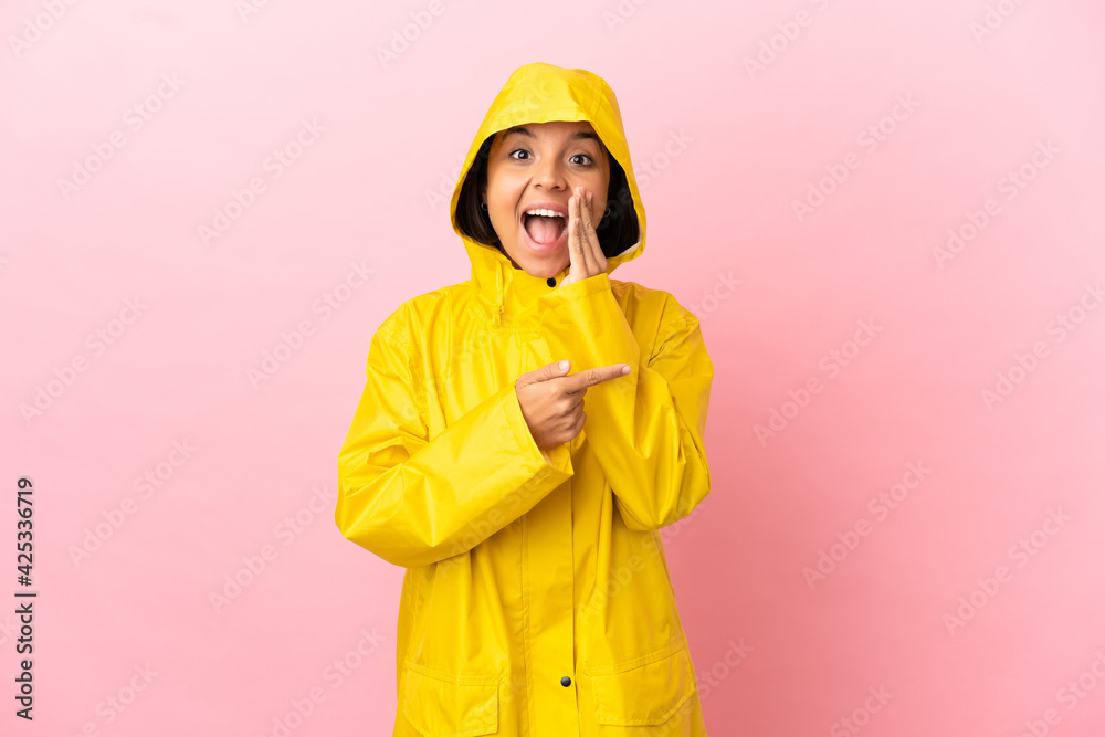 Young latin woman wearing a rainproof coat over isolated background pointing to the side to present a product and whispering something Young latin woman wearing a rainproof coat over isolated background pointing to the side to present a product and whispering something
