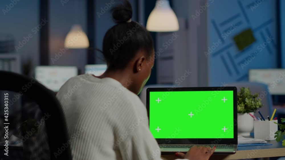 Dark skin freelancer working in front of greenscreen display of laptop sitting at desk in business office late at night. Businesswoman watching desktop with green mockup, chroma key, working overtime Dark skin freelancer working in front of greenscreen display of laptop sitting at desk in business office late at night. Businesswoman watching desktop with green mockup, chroma key, working overtime
