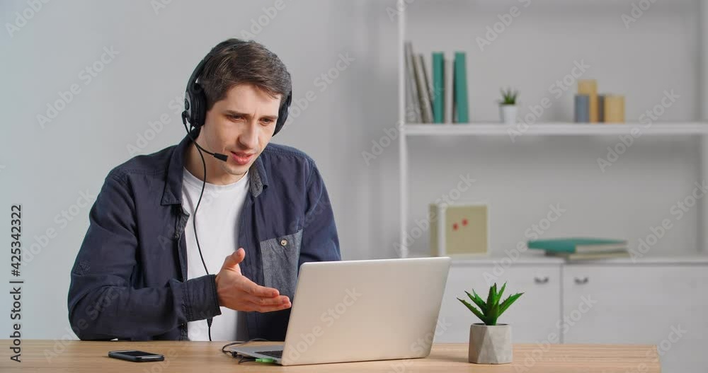 Business man in casual shirt sits at table makes video call chat wears headphones head microphone speaks into laptop web camera feels doubt negotiates speaking online shows thumbs up, remote talking Business man in casual shirt sits at table makes video call chat wears headphones head microphone speaks into laptop web camera feels doubt negotiates speaking online shows thumbs up, remote talking