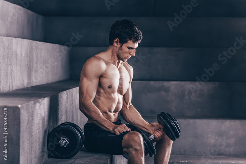 Strong muscle man sit and exercise biceps with heavy dumbbells.