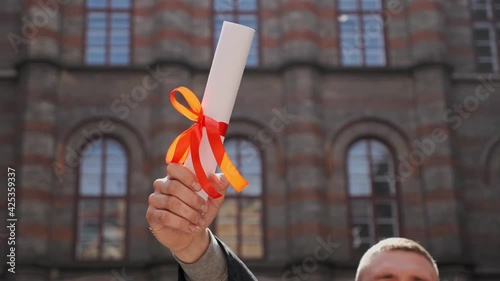 Male hand raised up holding diploma of academy graduate on the background of beautiful university school building Education concept Young graduated guy holding graduation degree convocation ceremony
