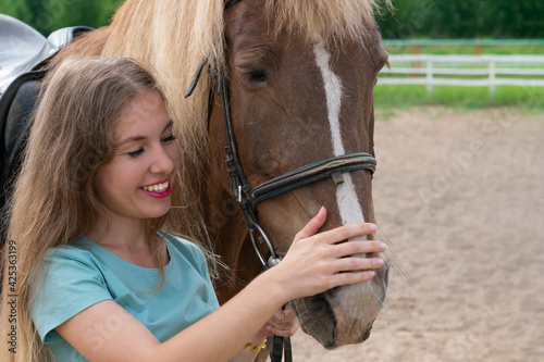 Portrait of young woman with blond long hair and smile is caressing brown horse snoot with big nostrils. Caring and breeding horses in rural ranch