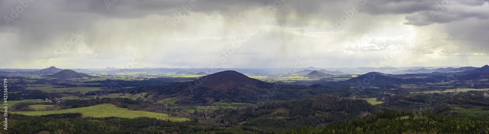 scenic view of rolling hills in the Bohemian Lusatian Mountains with patches of sunlight and storm clouds and rain plumes. Mount Ralsko, Mount Tlustek, Jezevcy vrch and Zeleny vrch can be seen