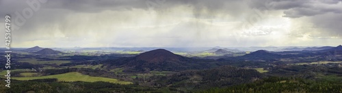 scenic view of rolling hills in the Bohemian Lusatian Mountains with patches of sunlight and storm clouds and rain plumes. Mount Ralsko, Mount Tlustek, Jezevcy vrch and Zeleny vrch can be seen photo
