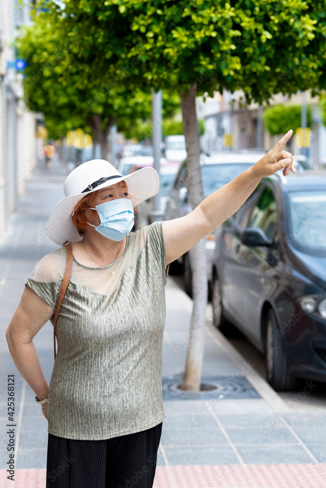 asian woman wearing a surgical mask and a hat pointing away outdoors. lifestyle concept.