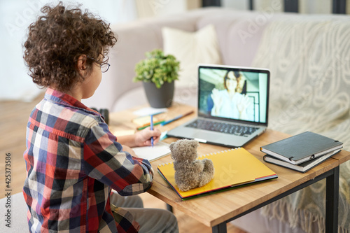 Get the knowledge. Rear view of hispanic school boy making notes during online lesson with teacher via video chat app. Child using laptop while studying at home