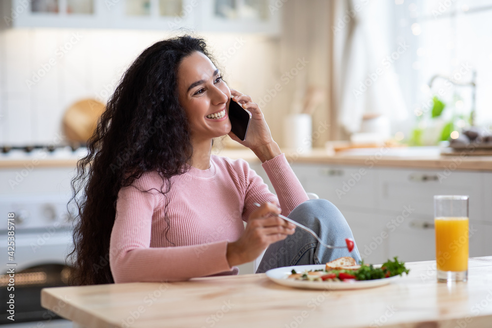 Smiling Millennial Lady Eating Breakfast And Talking On Phone In Kitchen