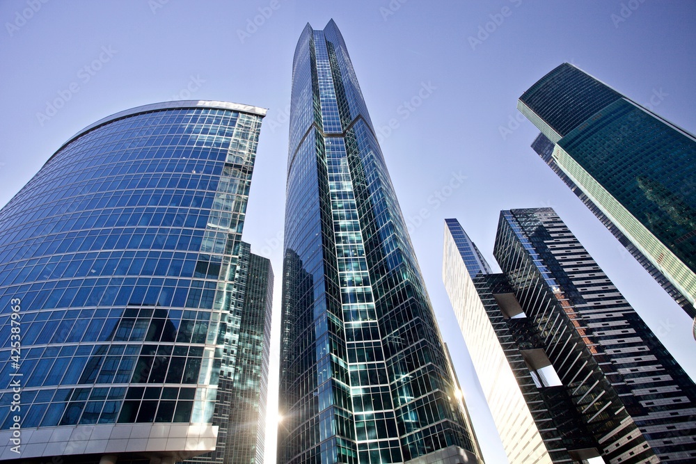 Obraz premium The bright sun shines through the tall modern skyscrapers. Office buildings against bright blue sky. Bottom-up view. Glass facades of tall skyscrapers with bright sun glare and reflections. Business.