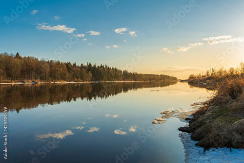 A deep blue river with reflective clouds and small pieces of ice. Forest and dry grass on the banks of the river. Spring landscape during sunset