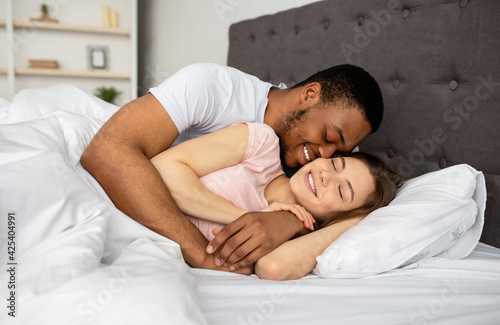 Obraz na plátně Multiracial couple cuddling in bed at home, hugging each other, having intimate