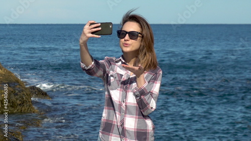 Young woman makes selfie on a smartphone on a background of the sea. Woman takes photos on vacation. Blue sea in sunny weather © Vital9c