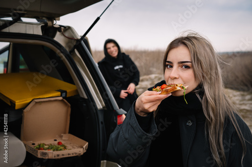 Woman and friend eating pizza slice by the car in winter trip