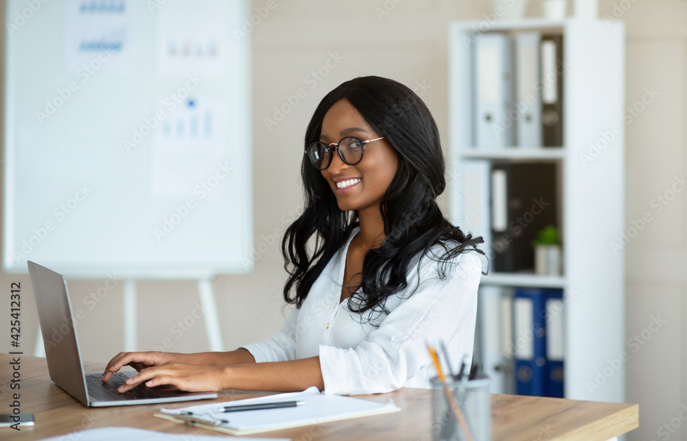 Happy young black businesswoman in glasses sitting at desk with open laptop at company office Happy young black businesswoman in glasses sitting at desk with open laptop at company office