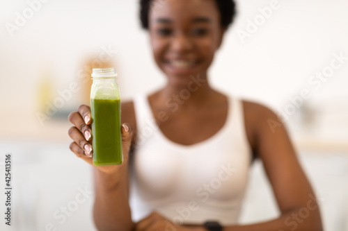 Slim African Female Showing Green Smoothie Bottle Sitting In Kitchen